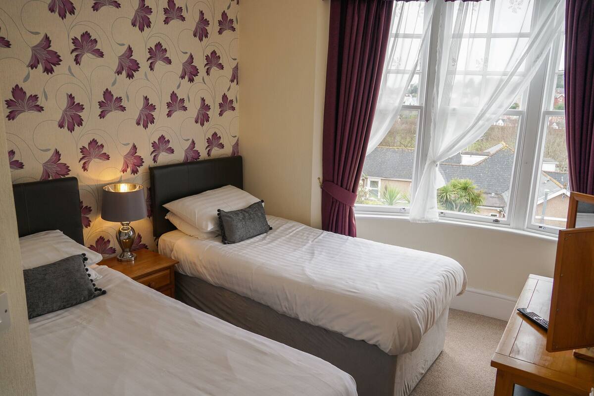 A hotel room with two single beds, a bedside table with a lamp, a window with curtains, and a view of the outside.