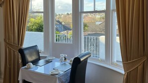 A dining area with a table set for two, large windows with curtains, and a view of a residential area. - The Sandpiper Guest House (Torquay)