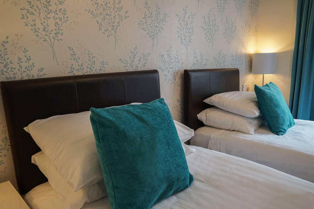 A hotel room with two beds, each with white bedding and teal pillows. The room has floral wallpaper and a bedside lamp.