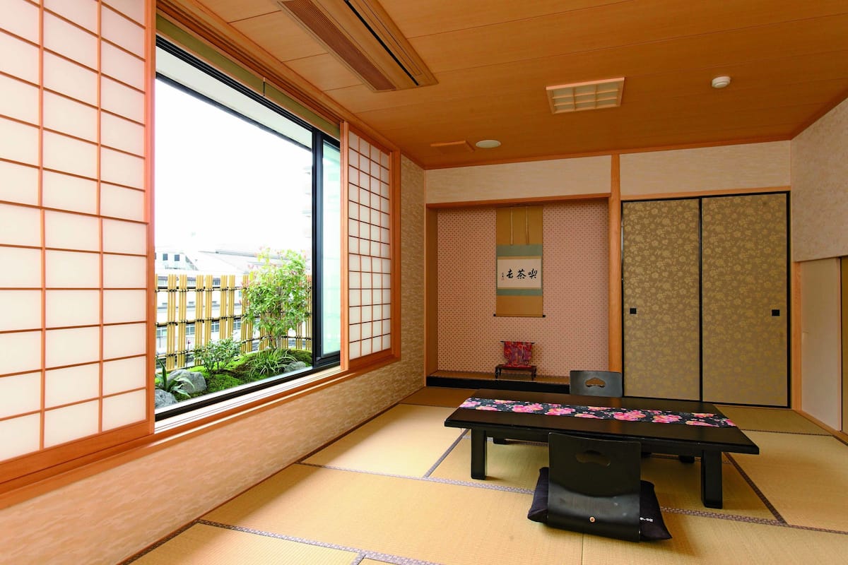 A traditional Japanese room with tatami flooring, a low table with a floral tablecloth, sliding doors, and a view of greenery outside.