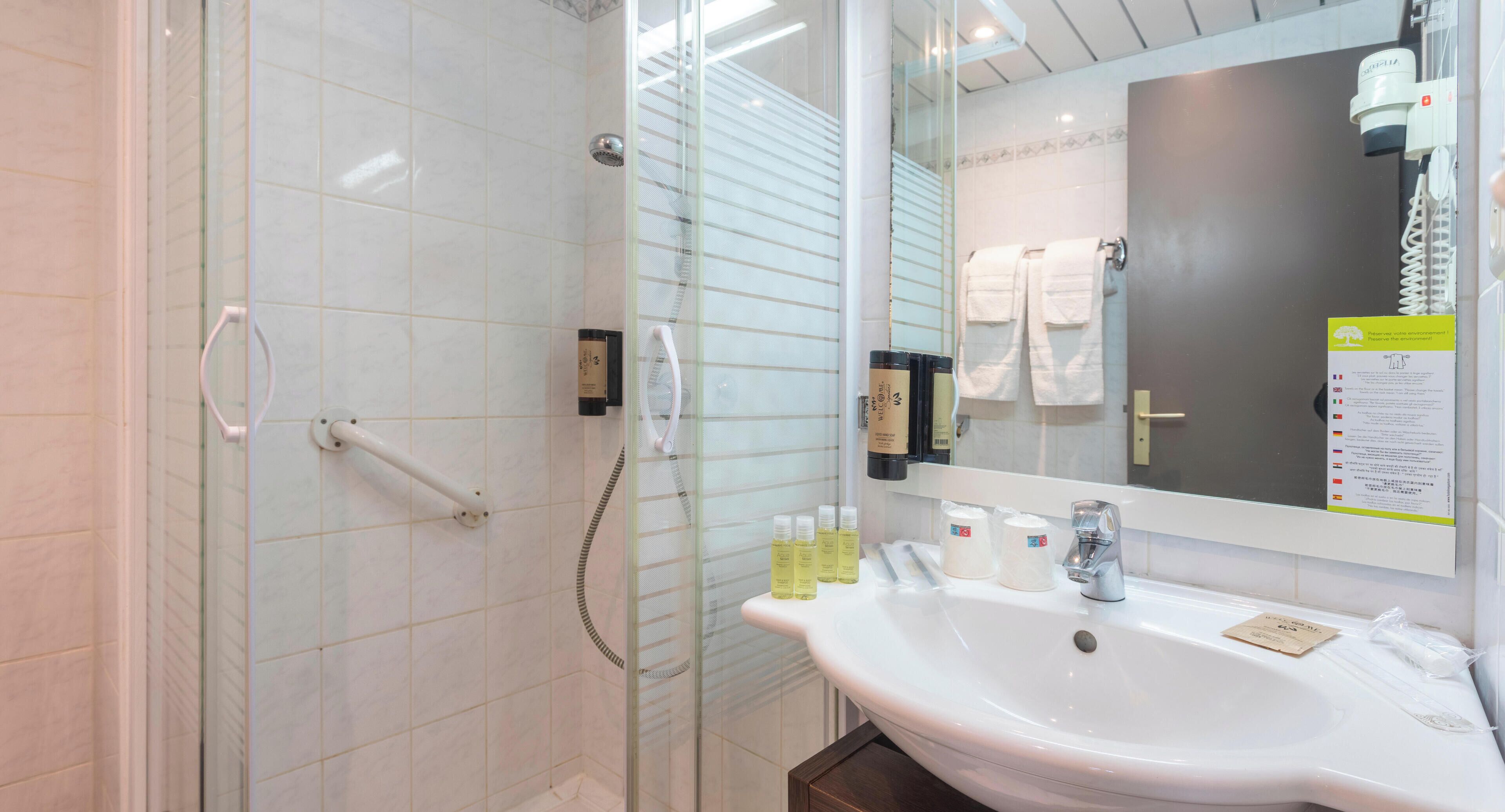 Une salle de bain moderne dotée d’une douche à parois de verre, d’un lavabo blanc avec des articles de toilette et d’un miroir muni d’un porte-serviettes.