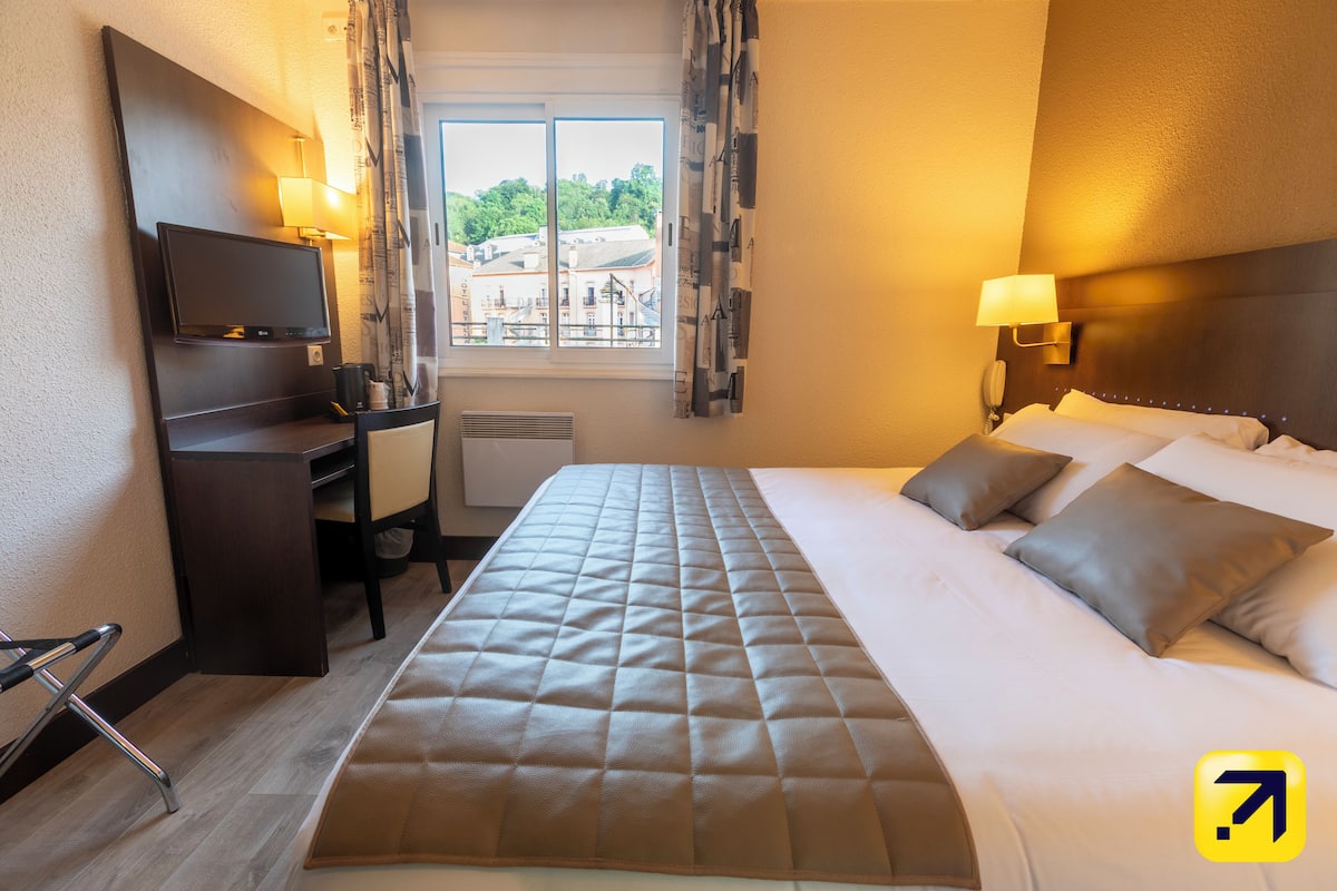 A hotel room with a bed, a desk, a chair, a TV, and a window with curtains.