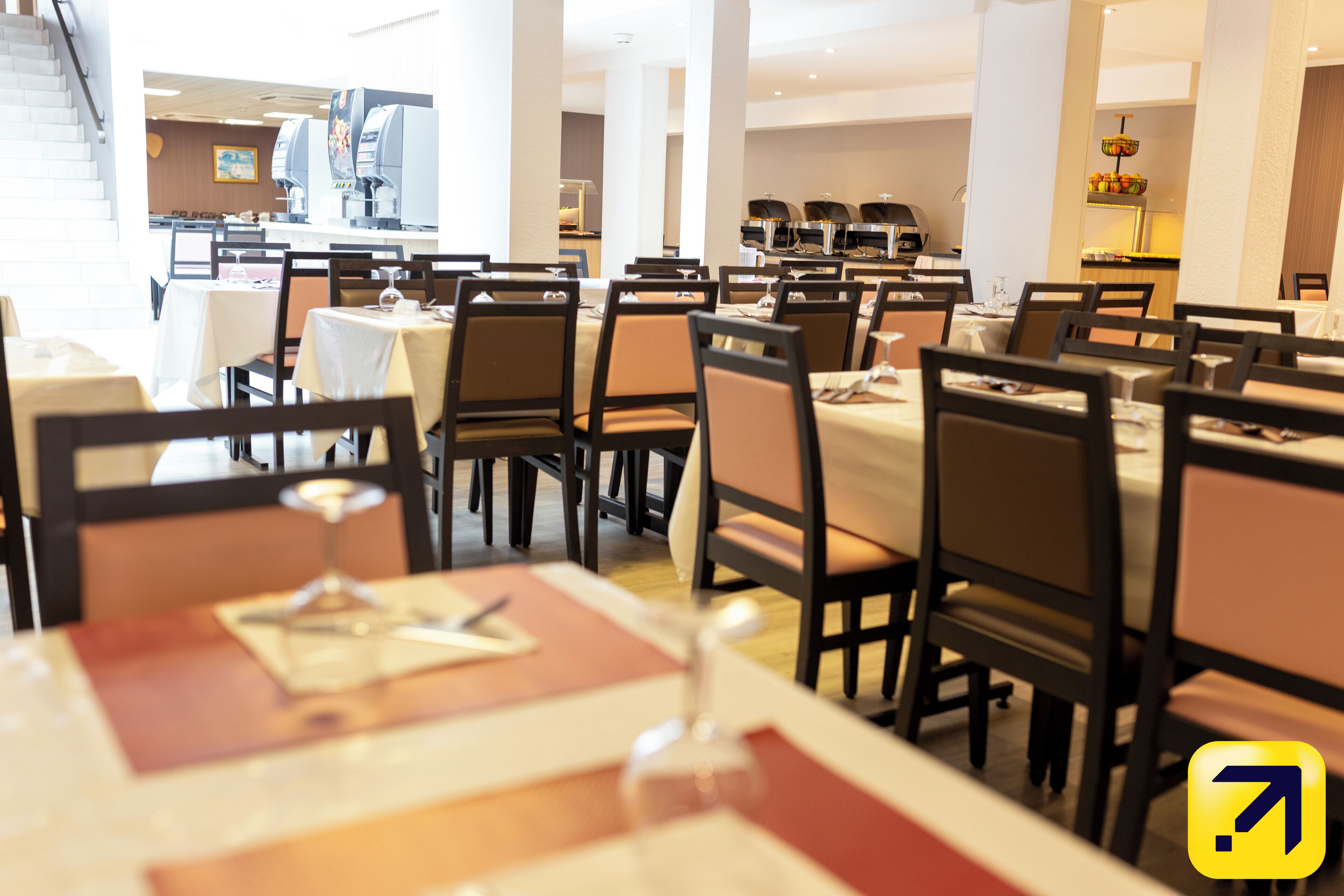 A dining area with tables set for service, chairs, and a buffet line in the background.