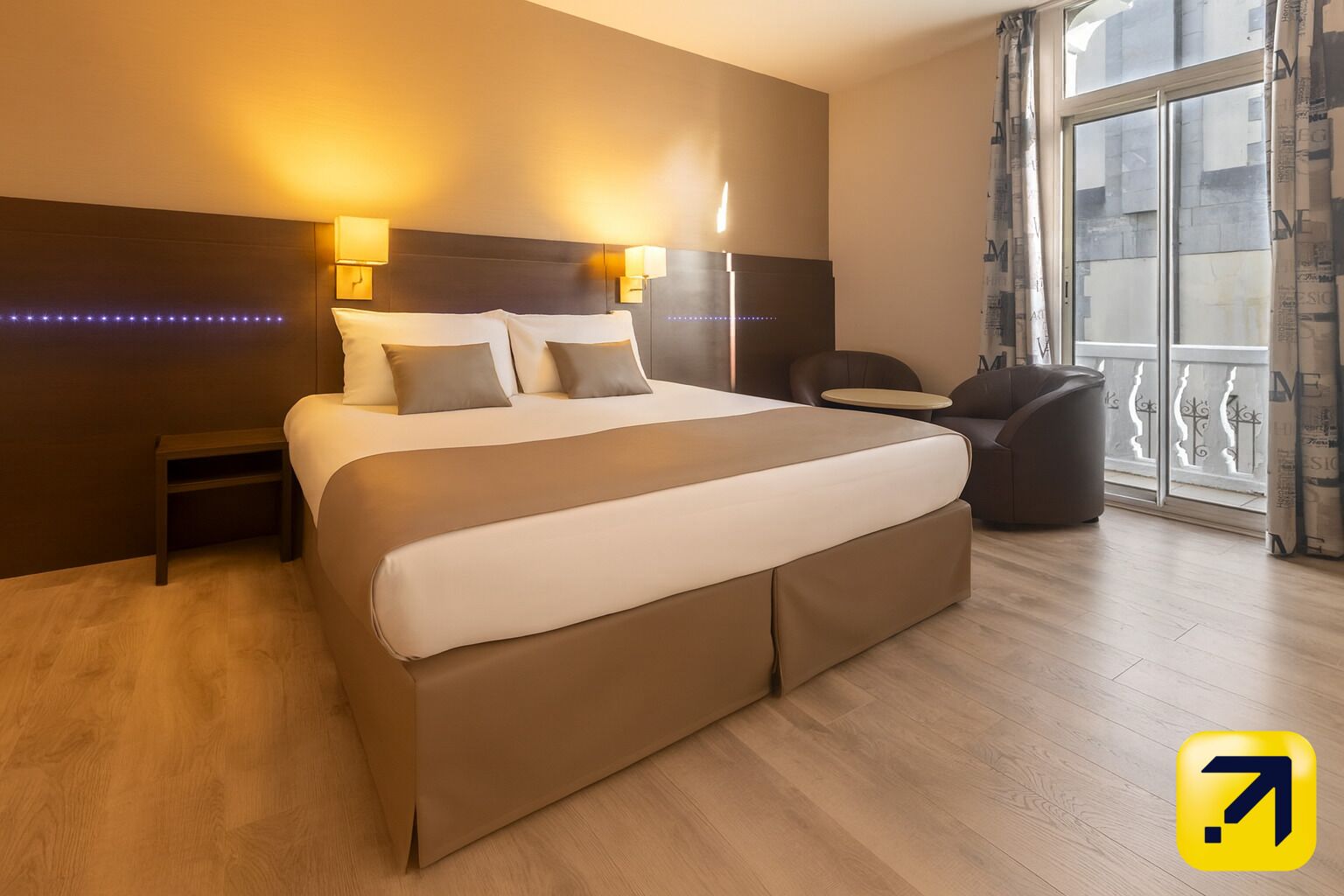 A modern hotel room with a large bed, bedside lamps, a small table, and a view of the city through large windows.