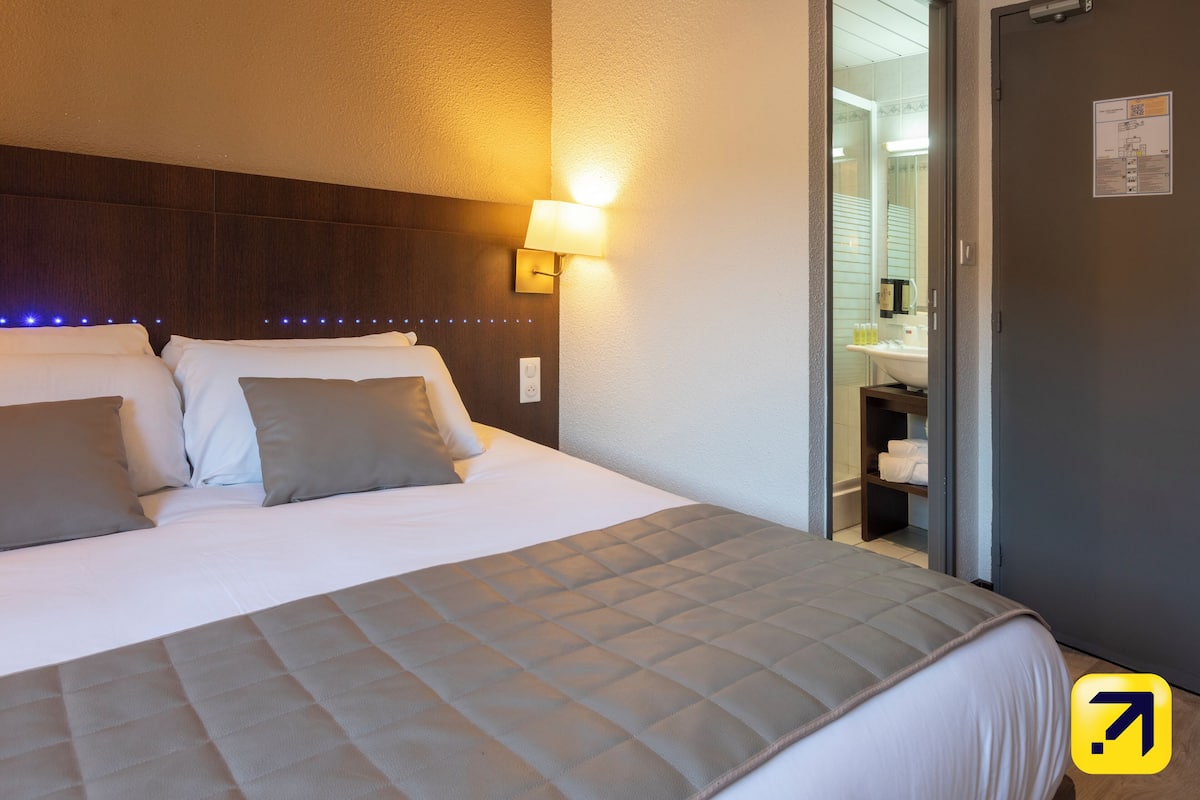 A hotel room with a bed, a headboard, a wall-mounted lamp, and a door leading to a bathroom.