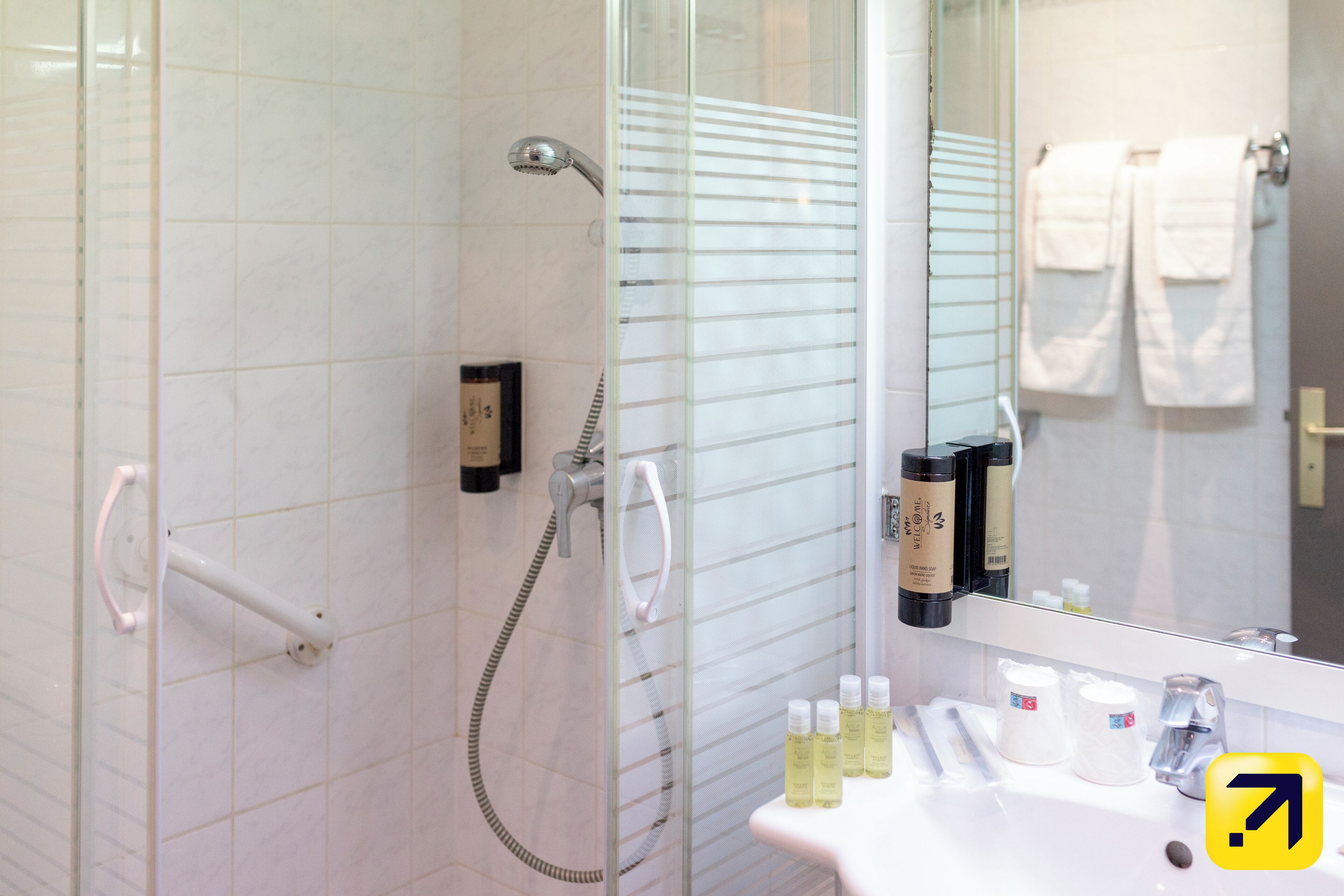 A modern bathroom with a glass shower enclosure, a white sink with toiletries, and a mirror reflecting a towel rack.