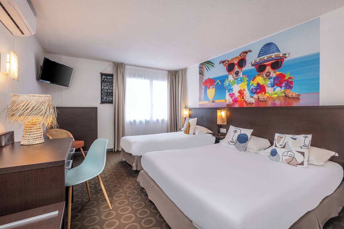 A hotel room with two beds, a desk, a chair, a TV, and a painting of dogs.