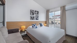 A hotel room with a large bed, a sofa, a small table with a lamp, and a view of the city through a large window. - The Original City, Hotel Cap Maya, Palais des congrès (Agde)