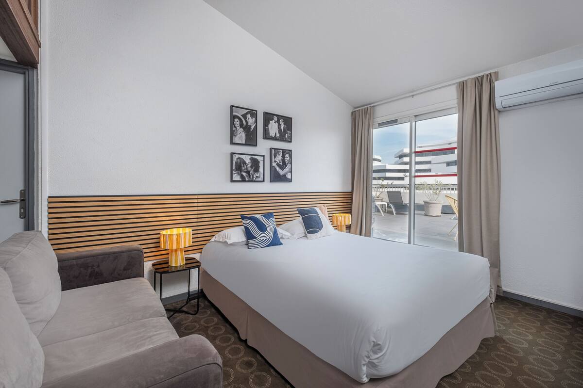 A hotel room with a large bed, a sofa, a small table with a lamp, and a view of the city through a large window.