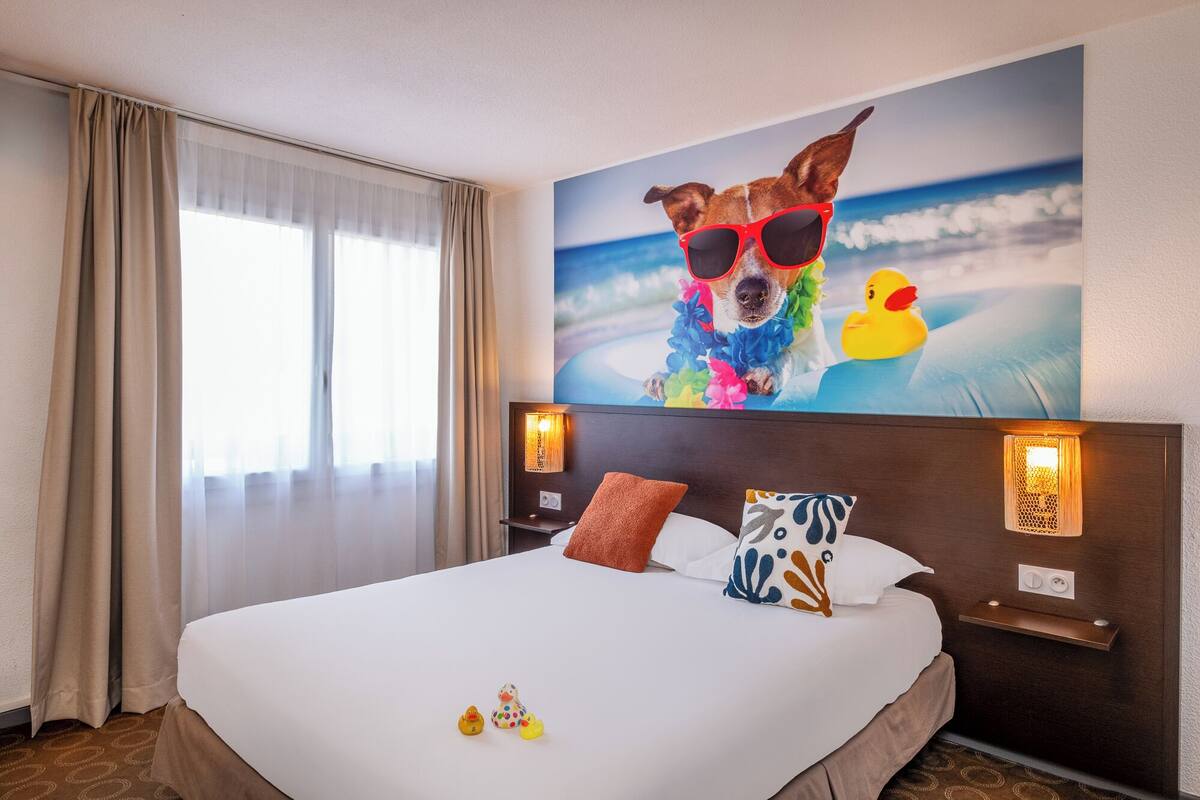 A hotel room with a large bed, a window with curtains, a wall-mounted artwork of a dog and a duck, and a nightstand with a lamp.