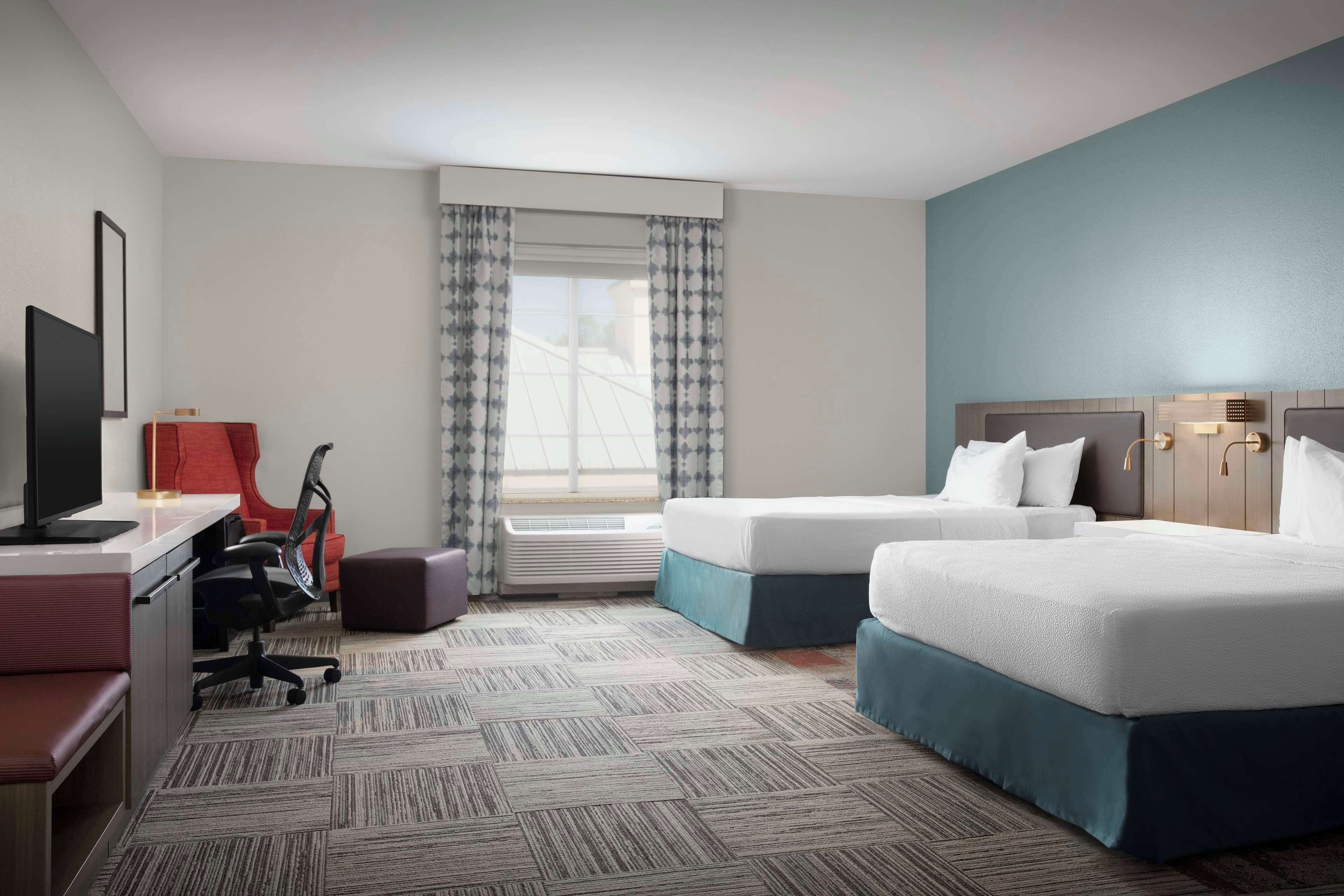 Een hotelkamer met twee bedden, een bureau met een computer, een stoel en een raam met gordijnen.