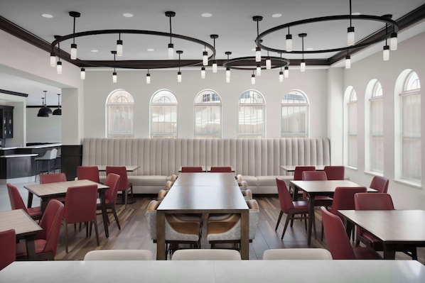 A modern dining area with a long table surrounded by red chairs, a sofa, and a kitchen in the background.