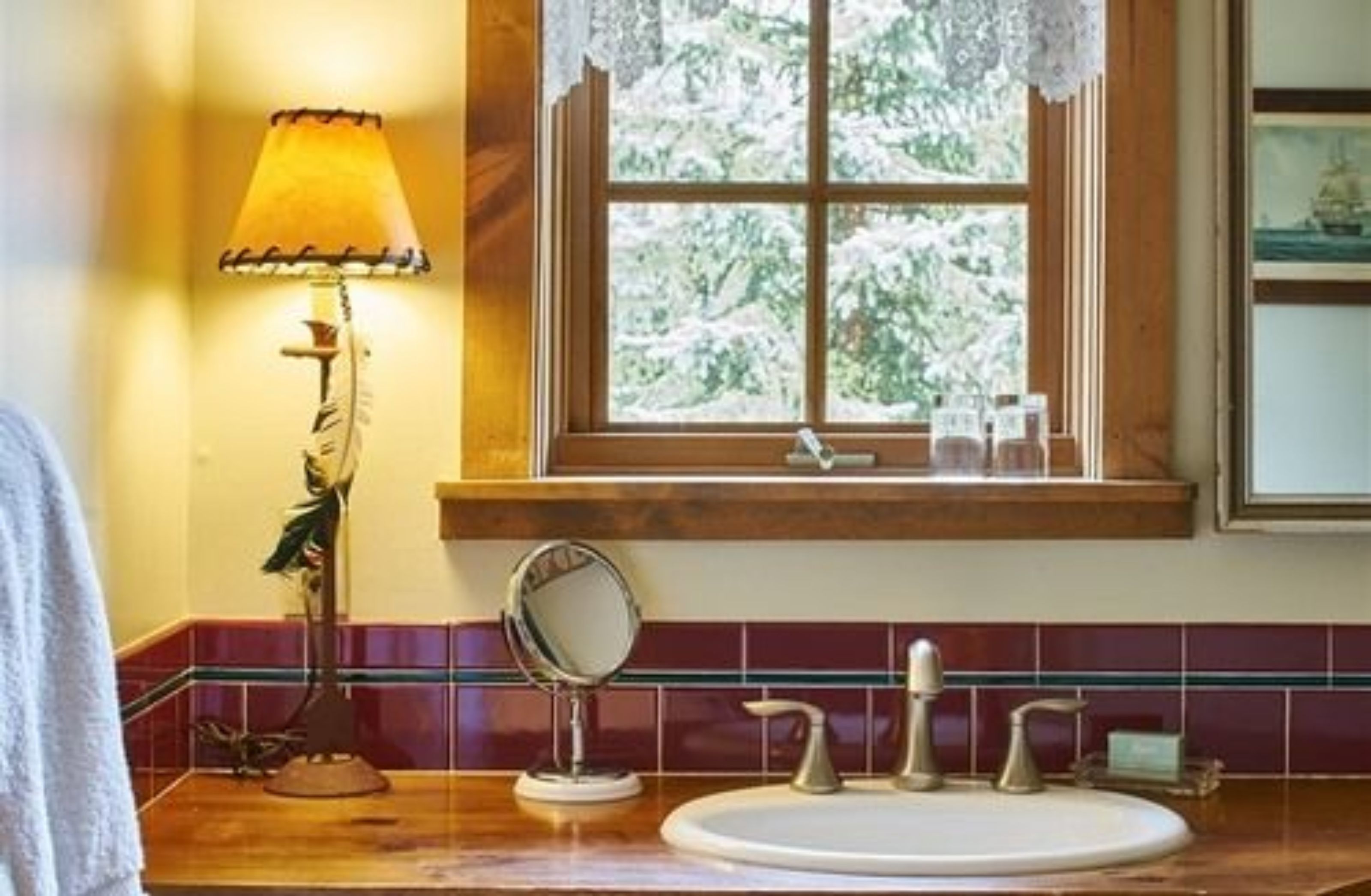 A bathroom with a wooden vanity, a round mirror, a sink with two faucets, and a window with a view of trees.