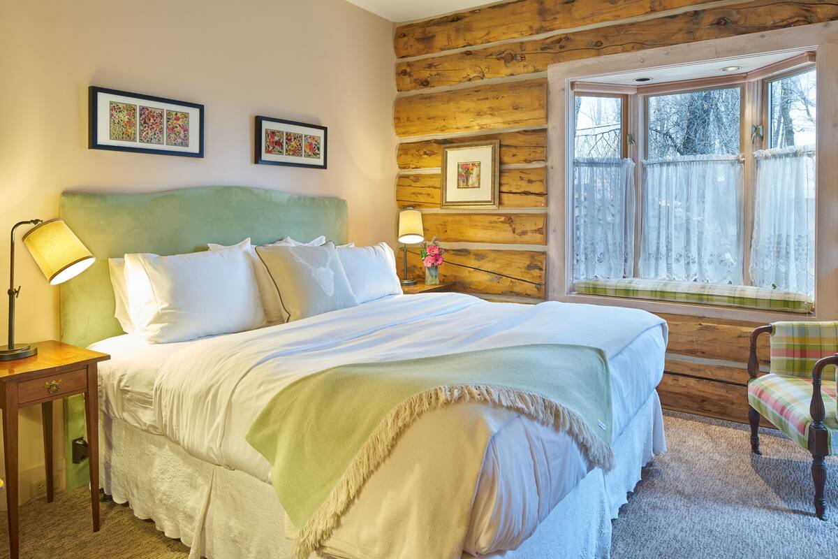 A cozy bedroom with a large bed, two bedside lamps, a chair, and a window with curtains.