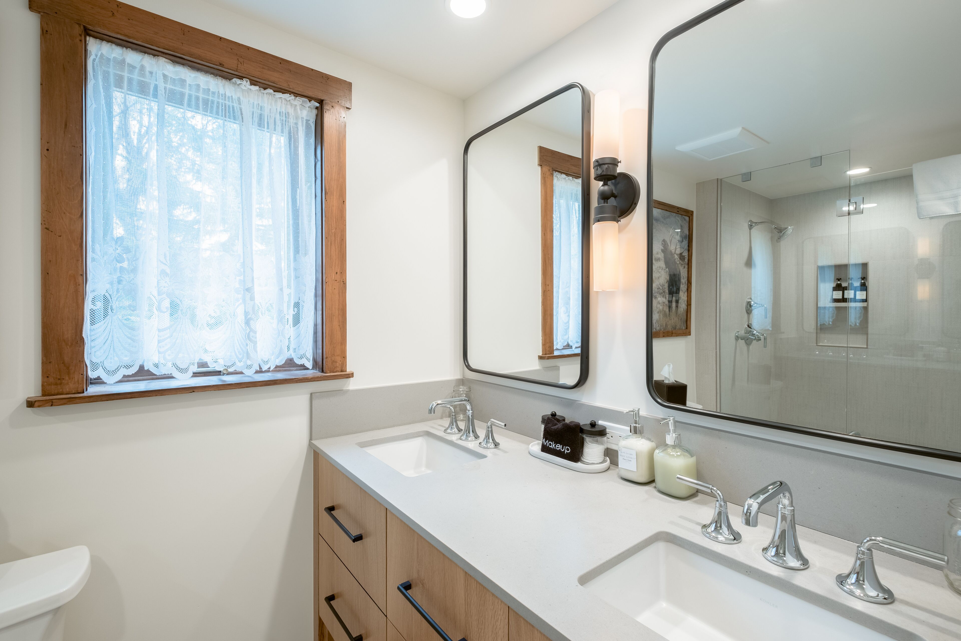 A bathroom with a double sink, a large mirror, and a window with lace curtains.