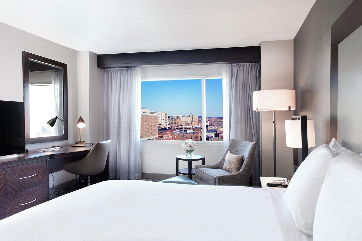 A hotel room with a large bed, a desk with a chair, a small table with a vase, and a view of the city through a large window.