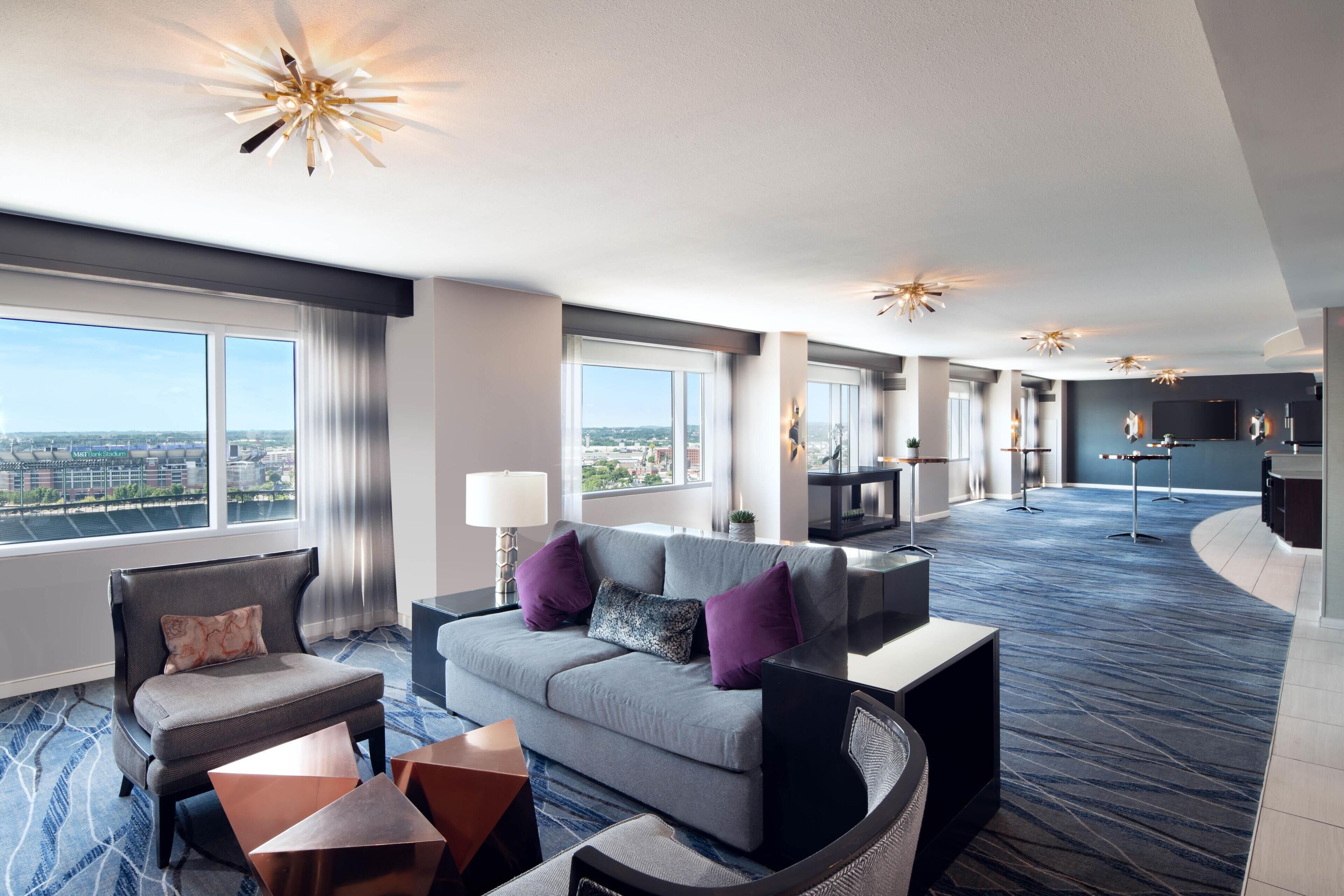 A modern hotel room with a sofa, armchair, and coffee table, large windows with a city view, and a dining area with a TV.