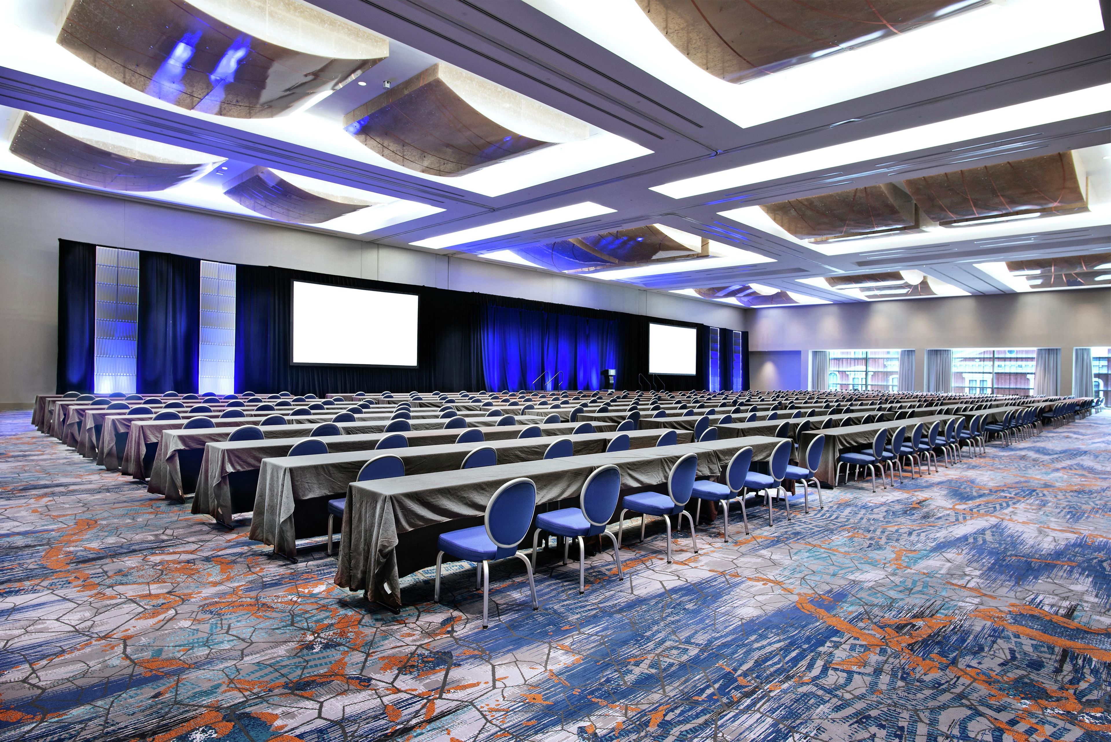A conference room with rows of tables and chairs, a large screen, and a patterned carpet.