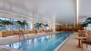 A large indoor swimming pool with lounge chairs and tables, surrounded by large windows and potted plants.