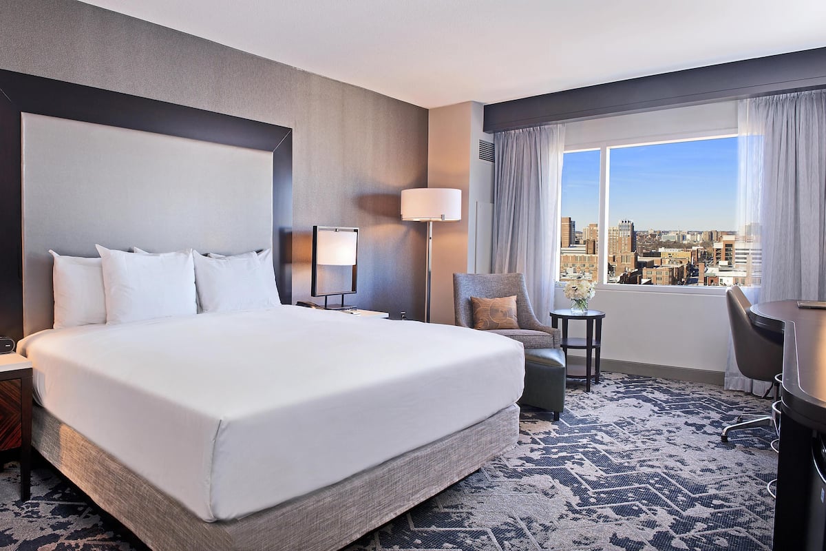 A hotel room with a large bed, a desk, a chair, a lamp, and a view of the city.