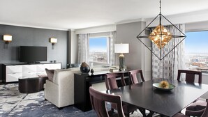 A modern hotel room with a flat-screen TV, a dining area with a table and chairs, a sofa, a decorative lamp, and a city view. - Hilton Baltimore Inner Harbor (Baltimore)