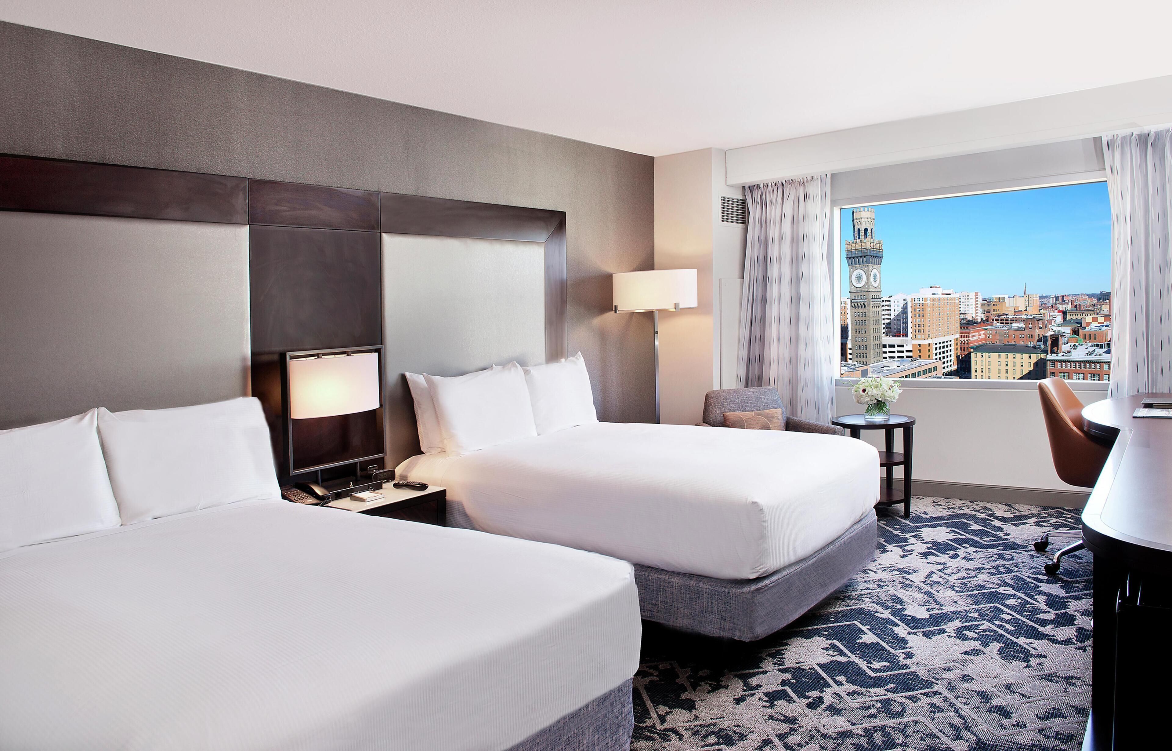 A hotel room with two beds, a desk, a lamp, a view of a cityscape, and a patterned rug.