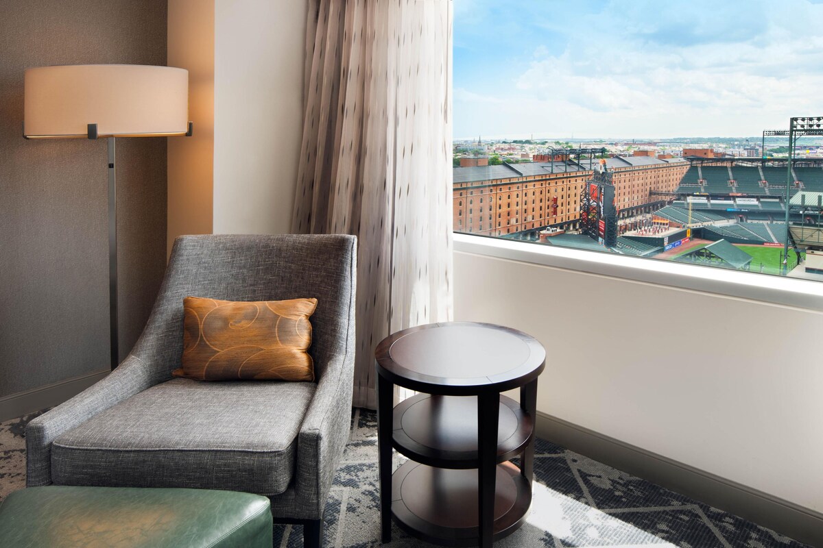A hotel room with a view of a stadium, a sofa, a small table, and a lamp.