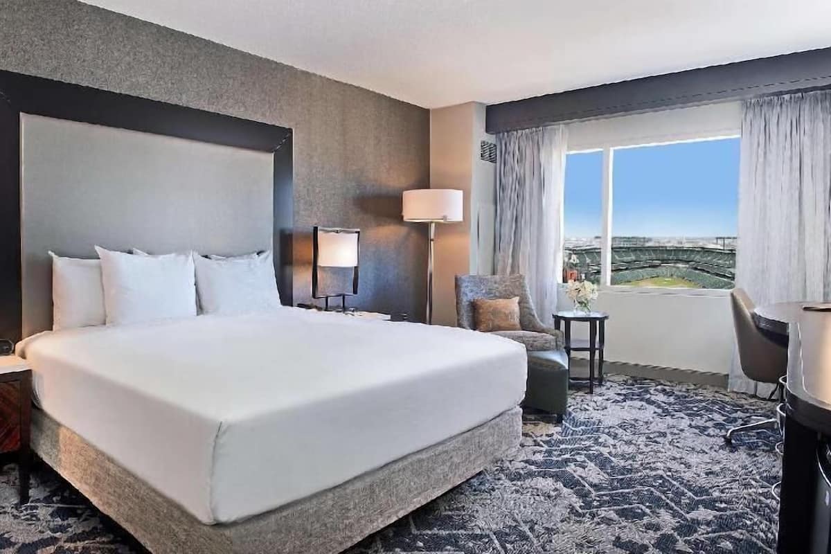 A hotel room with a large bed, a desk, a chair, a lamp, and a view of the city.