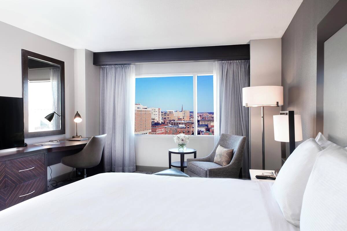 A hotel room with a large bed, a desk with a chair, a small table with a vase, and a view of the city through a large window.