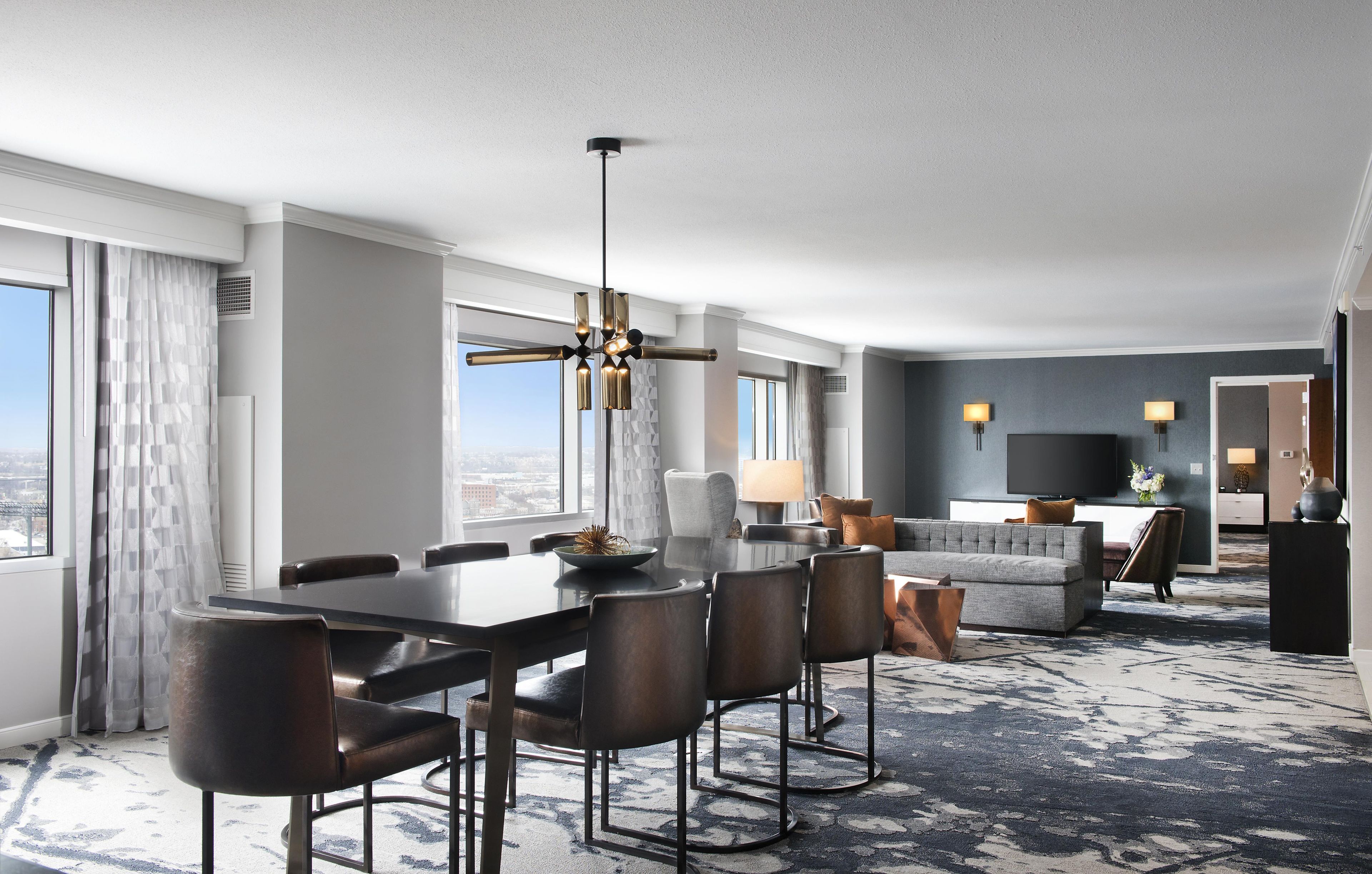 A modern hotel room with a dining area, a sofa, a TV, and a view of the city.