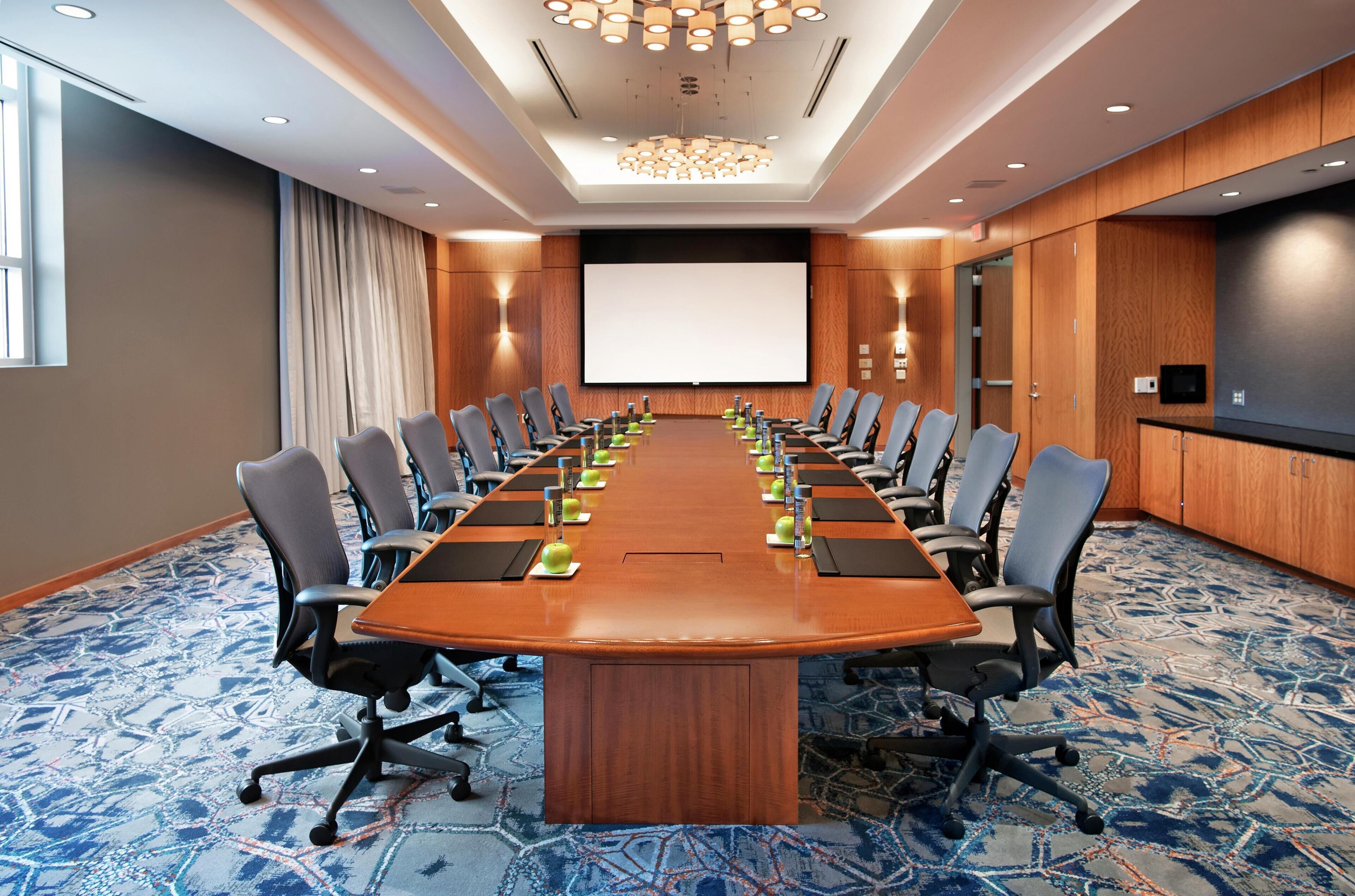 A conference room with a long table, chairs, a projector screen, and a carpeted floor.