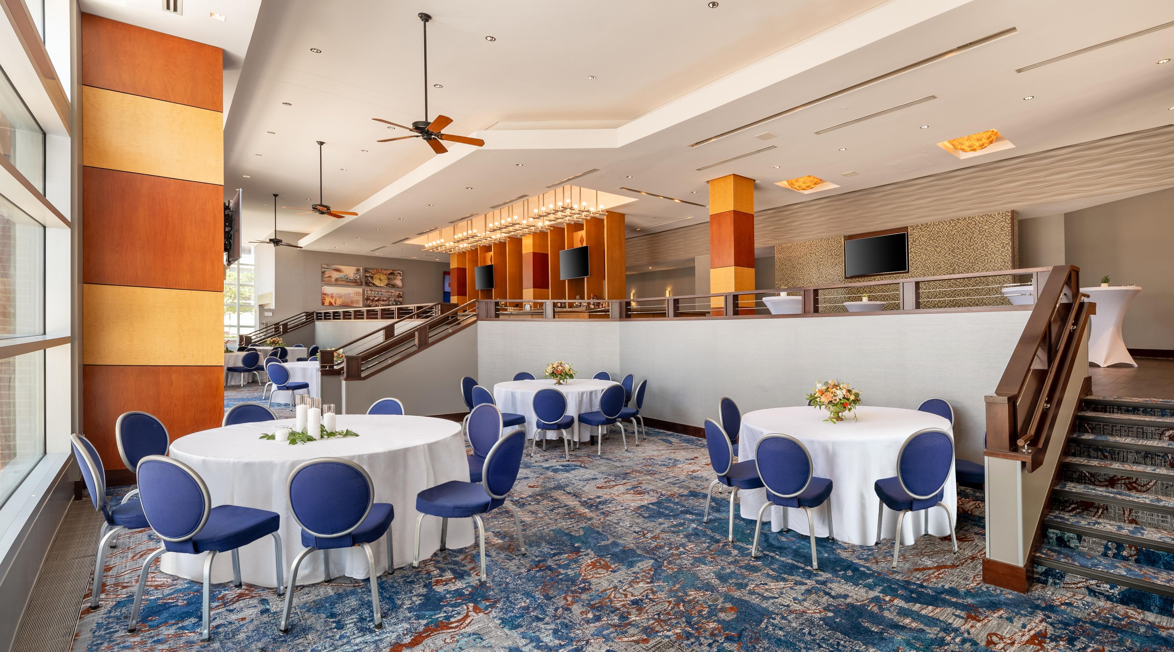 A hotel lobby with multiple round tables, blue chairs, a staircase, and a modern ceiling design.