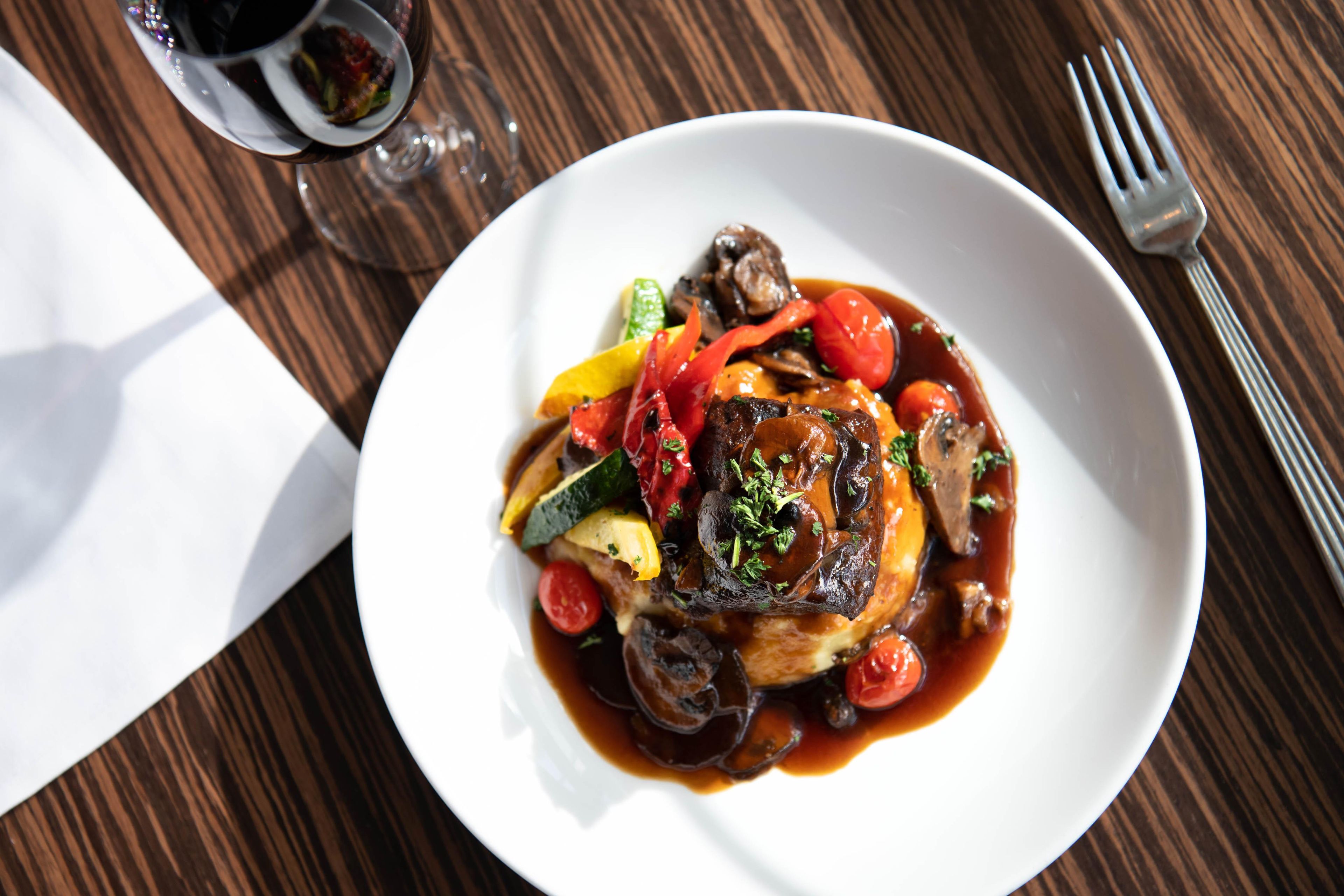 Ein Teller mit Essen, darunter verschiedene Gemüsesorten und Fleisch, dazu ein Glas Rotwein und eine Gabel auf einem Holztisch.