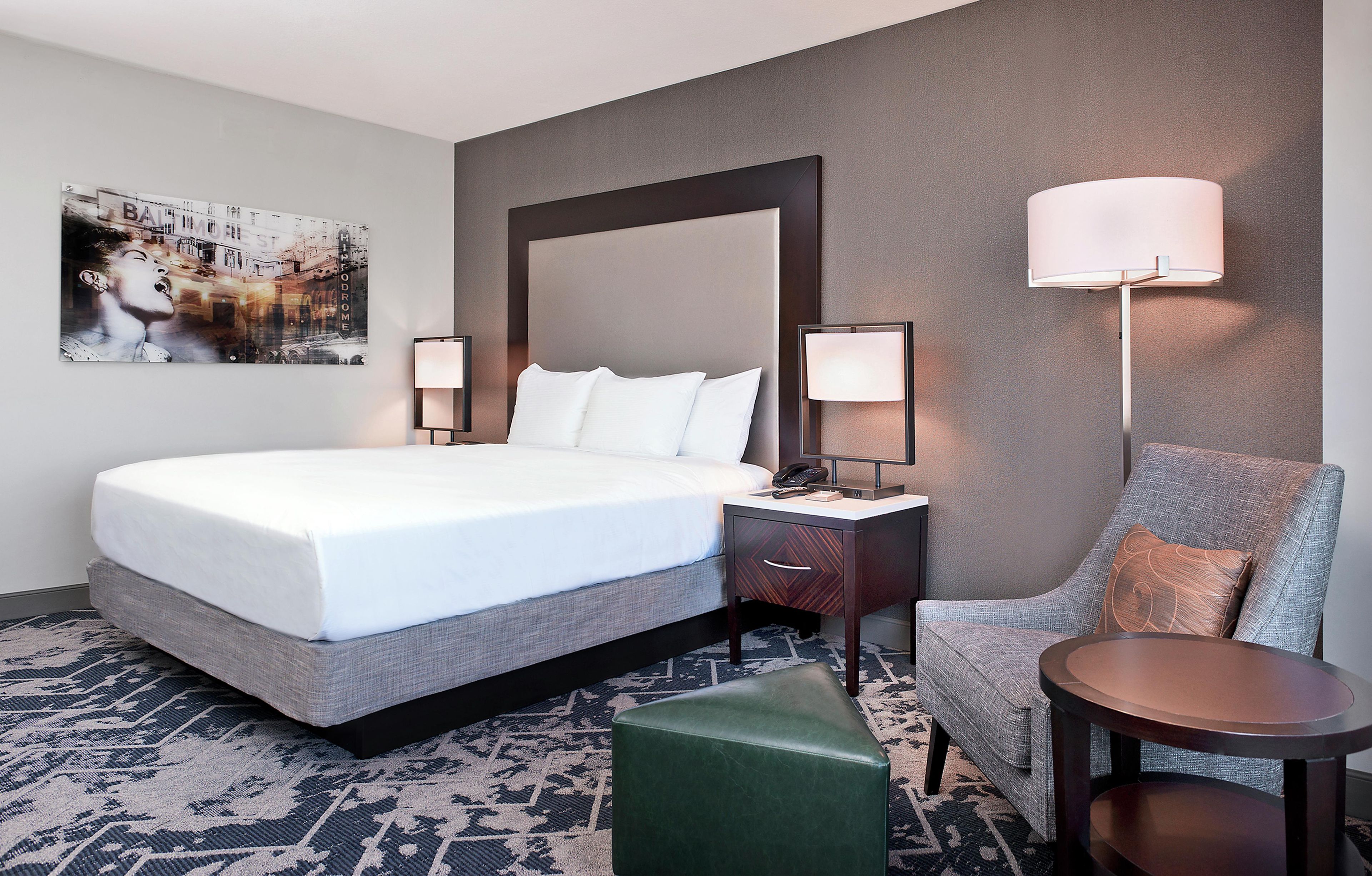 A hotel room with a large bed, bedside lamps, a chair, a small table, and a painting on the wall.