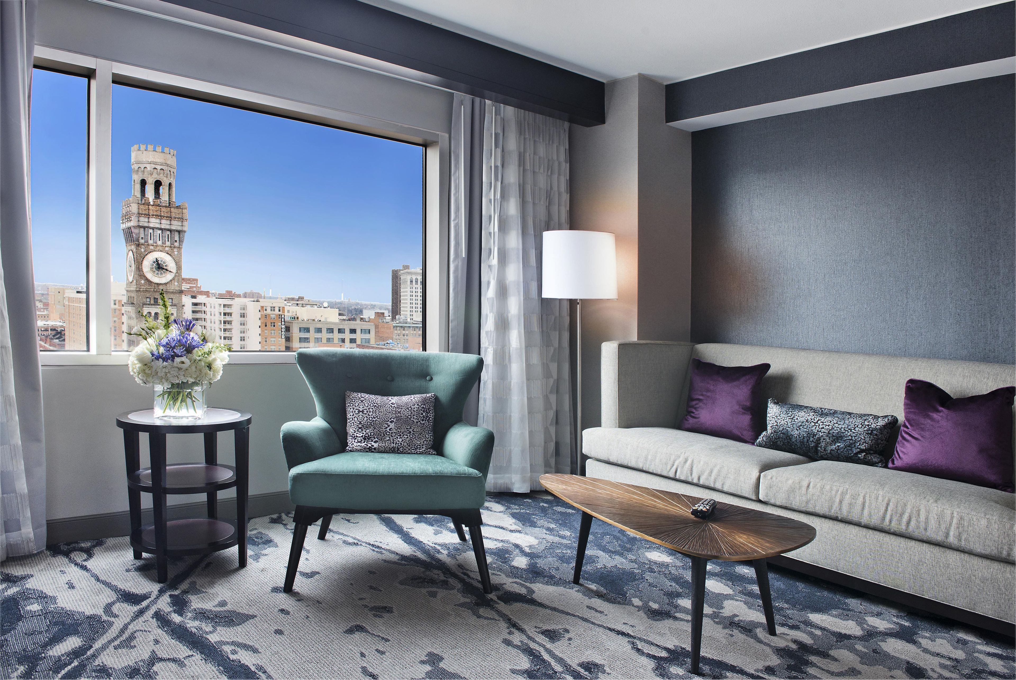 A hotel room with a view of a clock tower, a sofa, a chair, a lamp, and a small table.