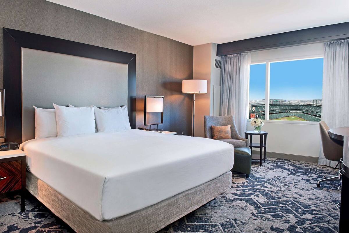 A hotel room with a large bed, a desk, a chair, a lamp, and a view of a stadium.