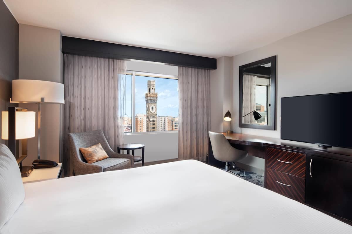 A hotel room with a large bed, a desk, a chair, a TV, a lamp, a phone, and a view of a clock tower.