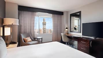 A hotel room with a large bed, a desk, a chair, a TV, a lamp, a phone, and a view of a clock tower.