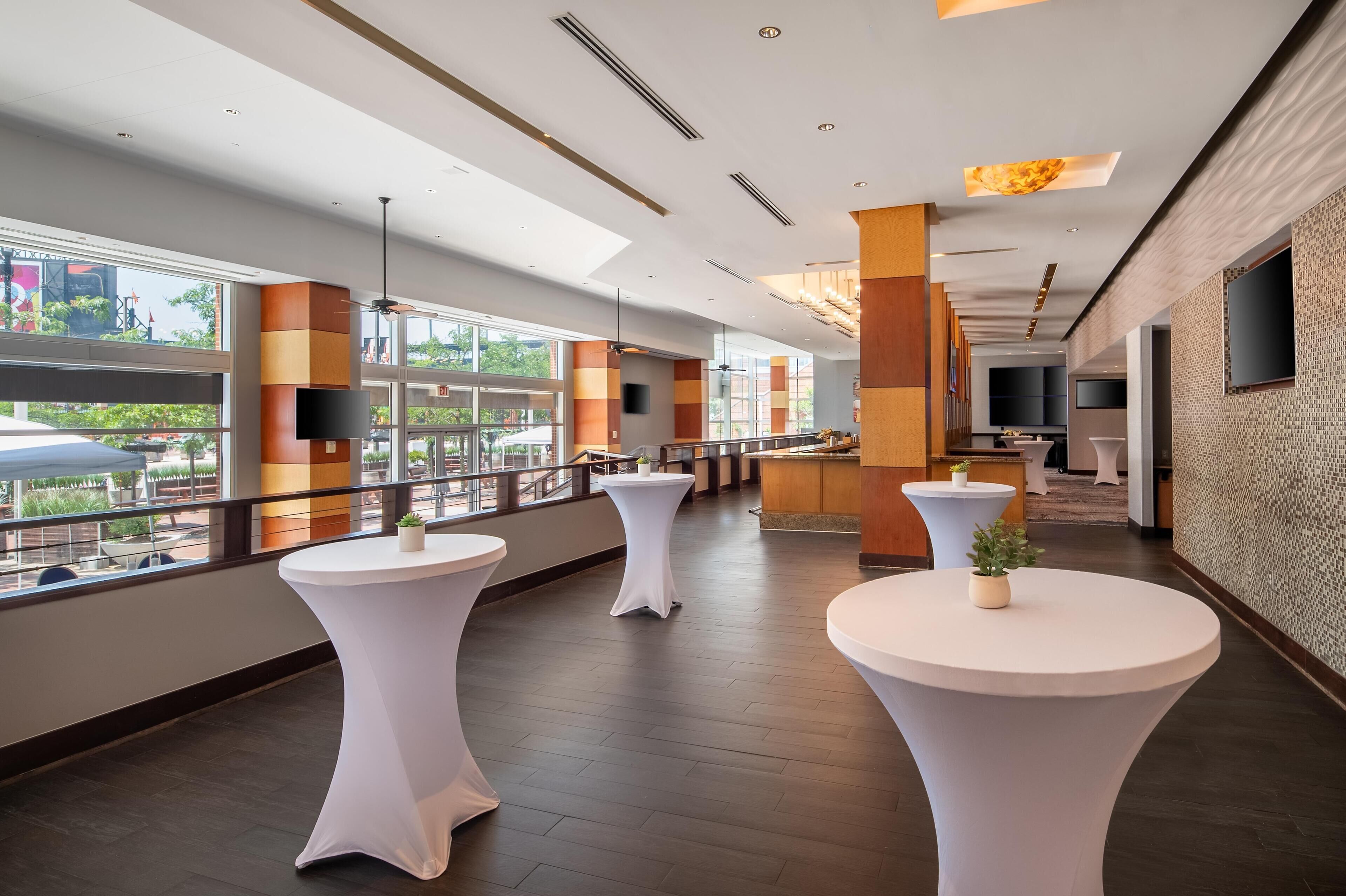 A modern hotel lobby with large windows, white round tables, and a flat-screen TV mounted on the wall.