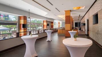 A modern hotel lobby with large windows, white round tables, and a flat-screen TV mounted on the wall.