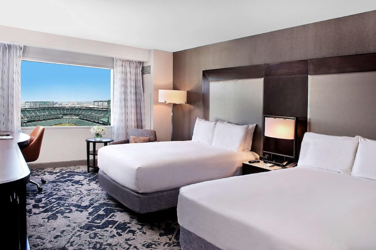 A hotel room with two beds, a desk, a chair, a lamp, and a view of a stadium.