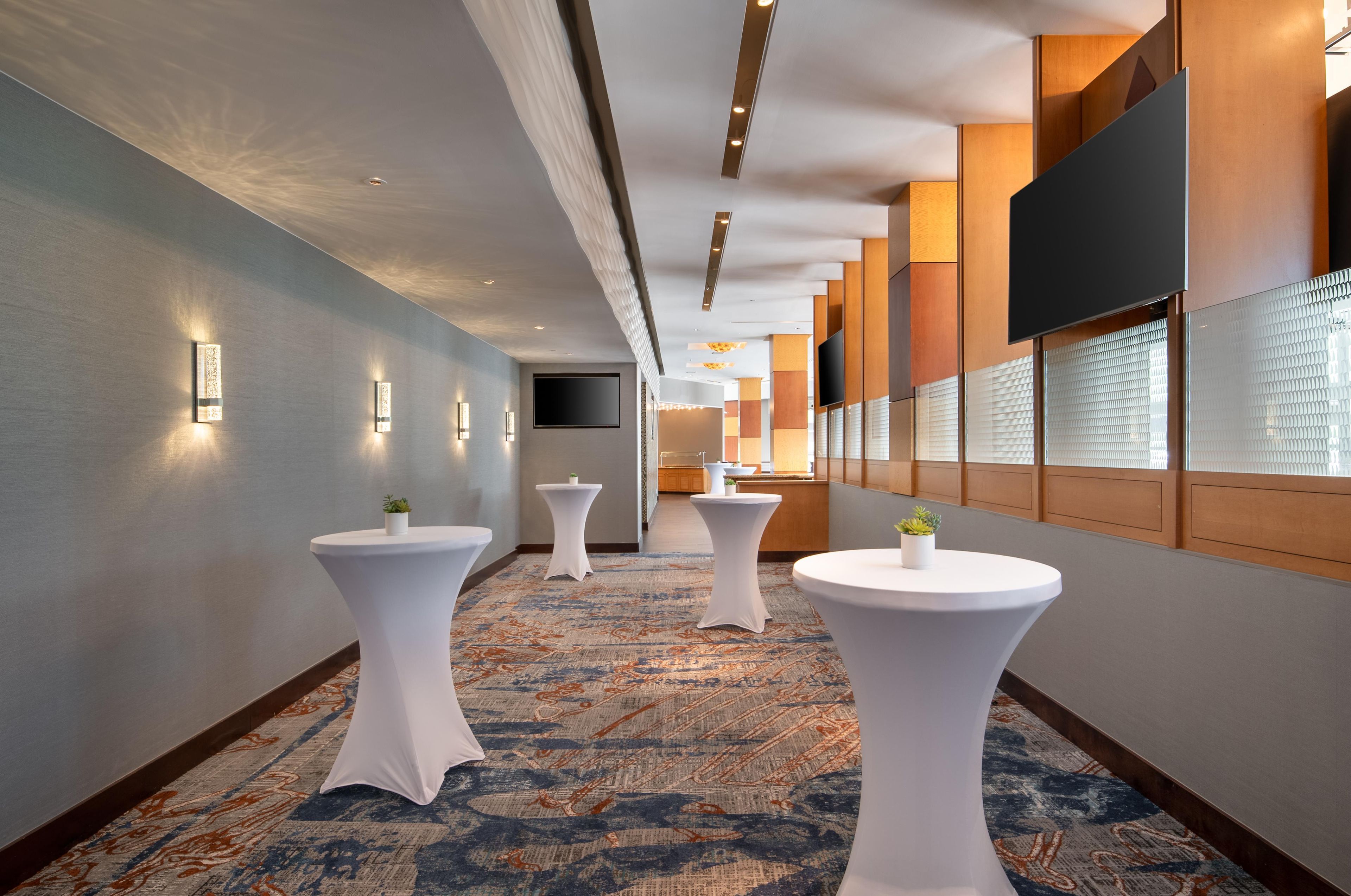 Un couloir d’hôtel avec des tables rondes blanches, un téléviseur à écran plat fixé au mur et une moquette à motifs.