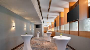Un couloir d’hôtel avec des tables rondes blanches, un téléviseur à écran plat fixé au mur et une moquette à motifs.