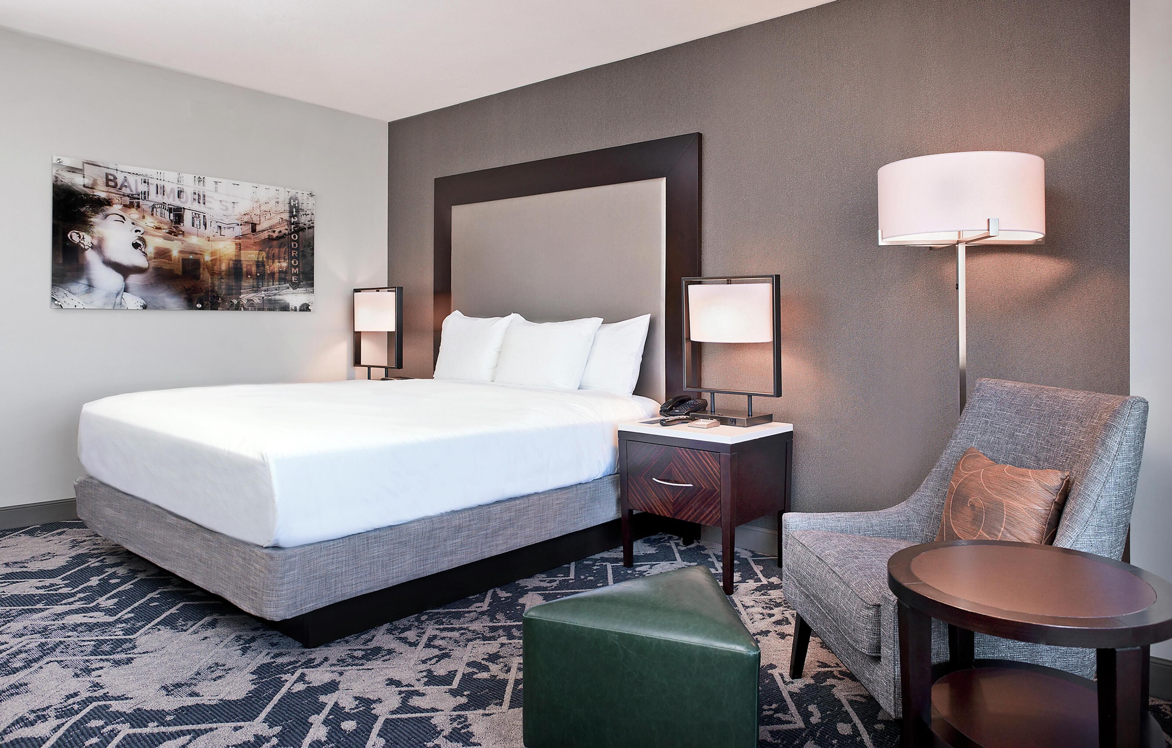A hotel room with a large bed, bedside lamps, a chair, a small table, and a painting on the wall.