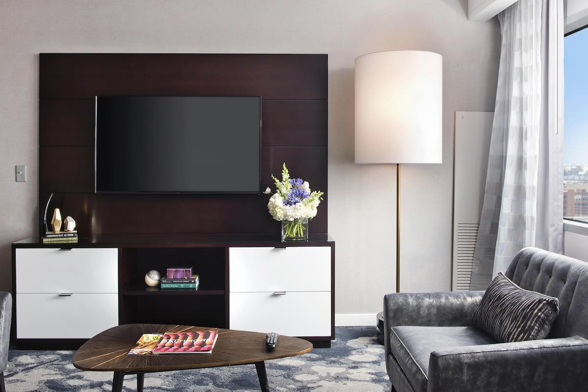 A modern hotel room with a flat-screen TV mounted on a dark wood panel, a white console with drawers, a grey sofa, a coffee table with a remote control, and a vase of flowers.