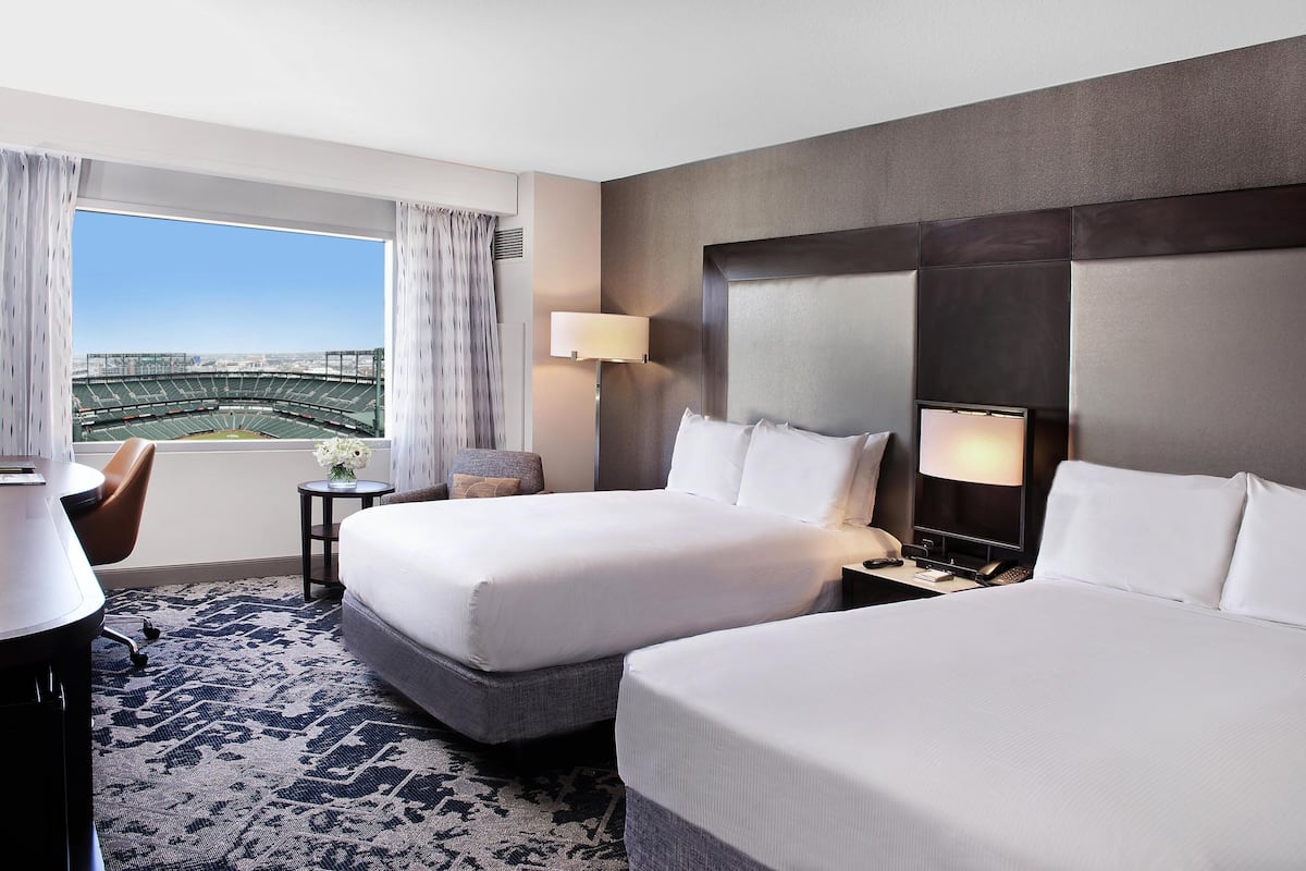 A hotel room with two beds, a desk, a chair, a small table with a lamp, and a view of a stadium.