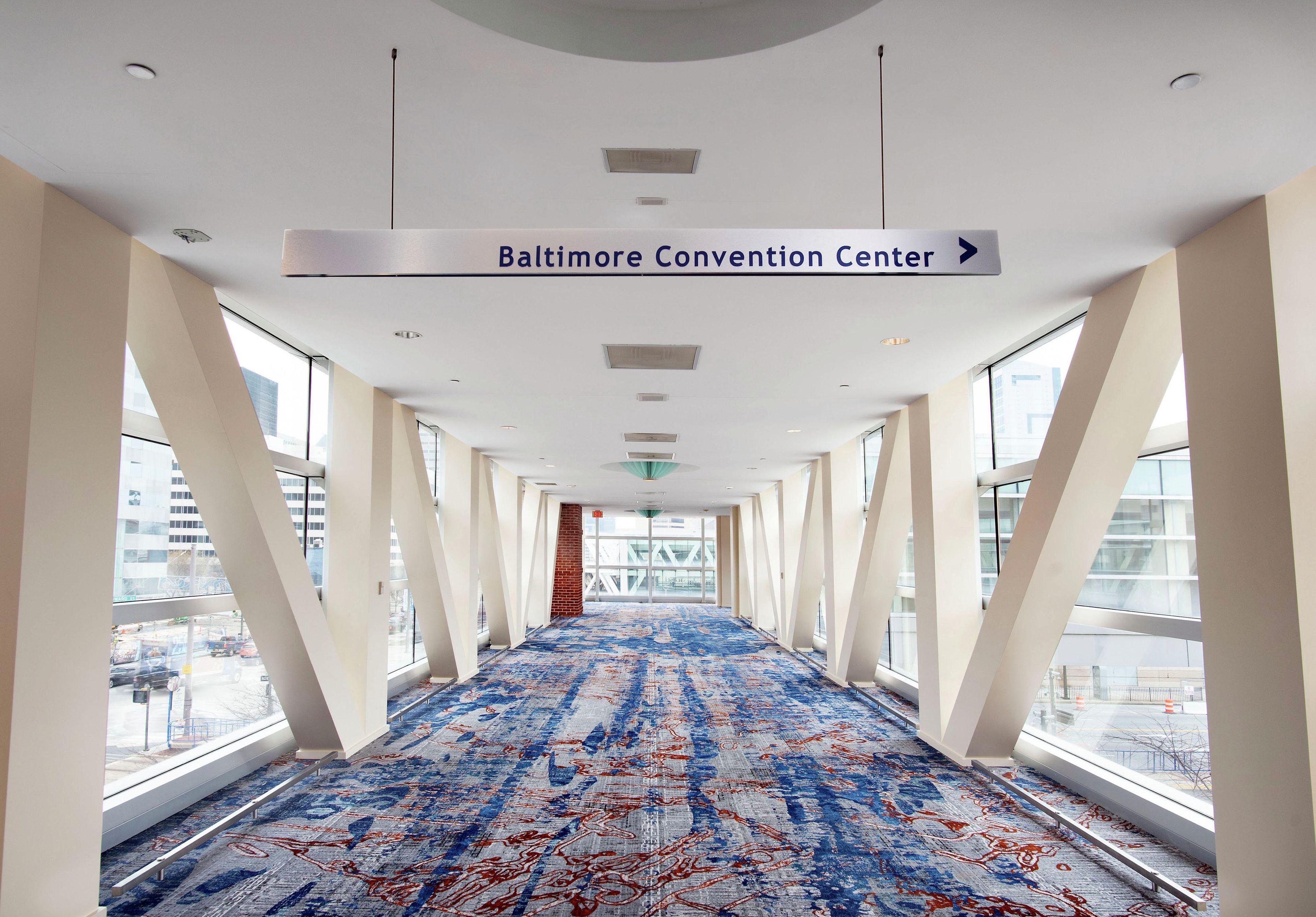 Un pasillo con un cartel indicativo hacia el Centro de Convenciones de Baltimore, amplias ventanas y una alfombra estampada.