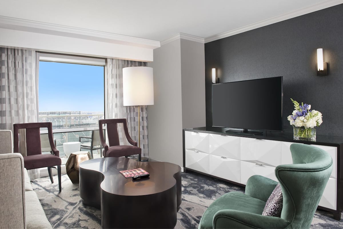 A hotel room with a large TV, two chairs, a sofa, a coffee table, and a view of the city.