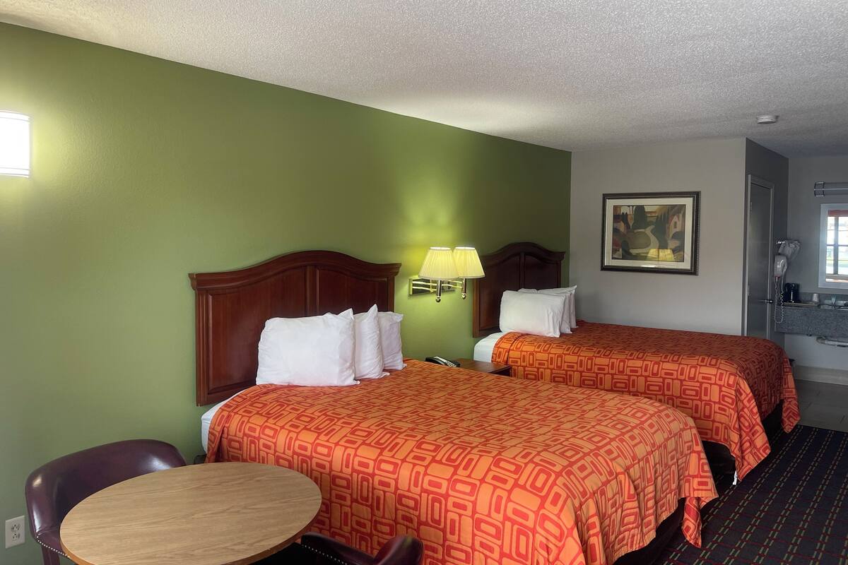 A hotel room with two beds, a round table, a chair, a lamp, and a painting on the wall.