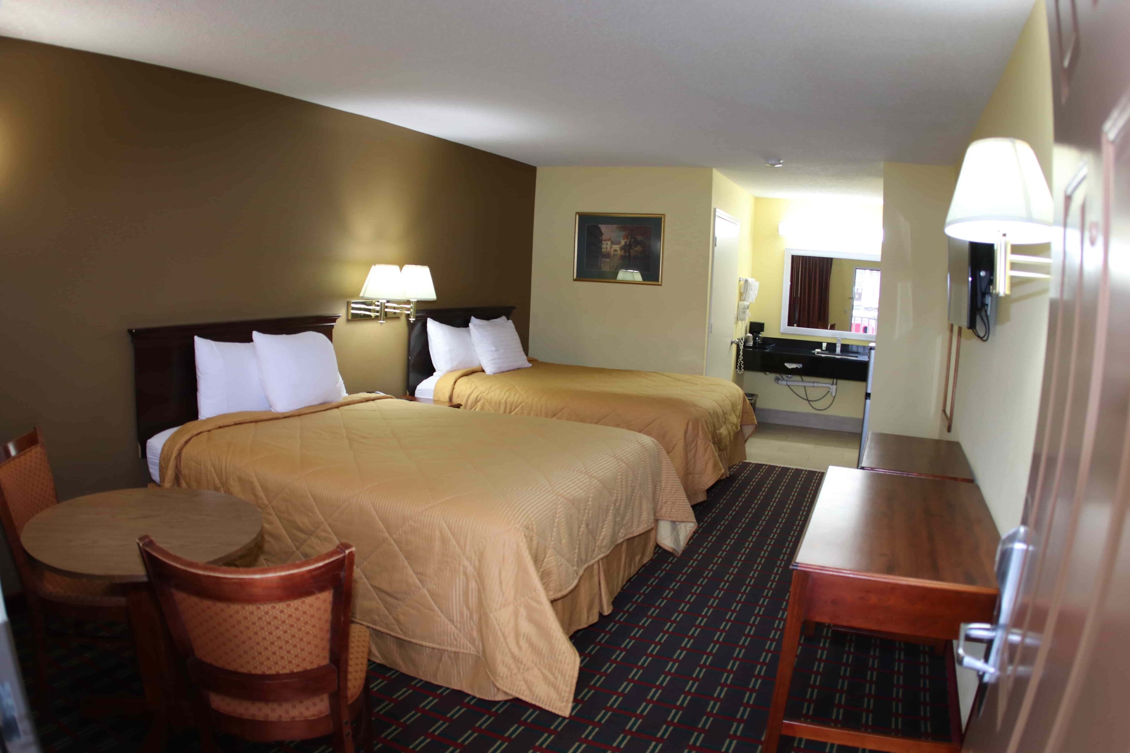 A hotel room with two beds, a desk, a chair, a table, a mirror, and a bathroom visible in the background.
